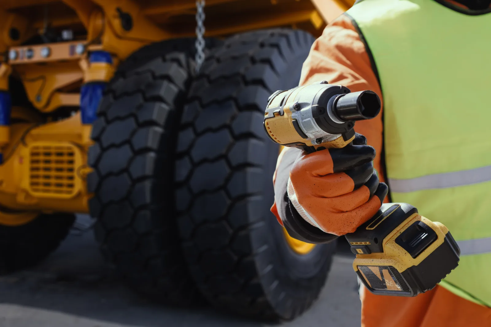Maintenance worker in high-visibility clothing and gloves holding an impact wrench beside a heavy-vehicle tyre.